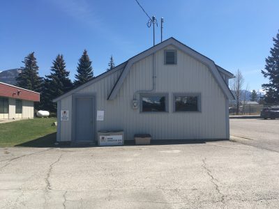 A photo of the Book Donation Centre. A grey shed with a bin out front. Blue sky behind. This donation centre is located next to the Invermere Courthouse.