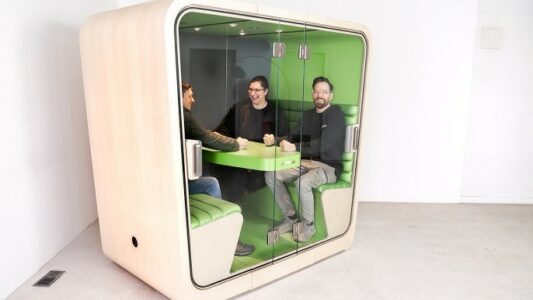 Loop booth with three people sitting inside. A cube-shaped pod with booth seating on either side of a table in the middle. Glass doors to get in and out and provide a soundproof space. 